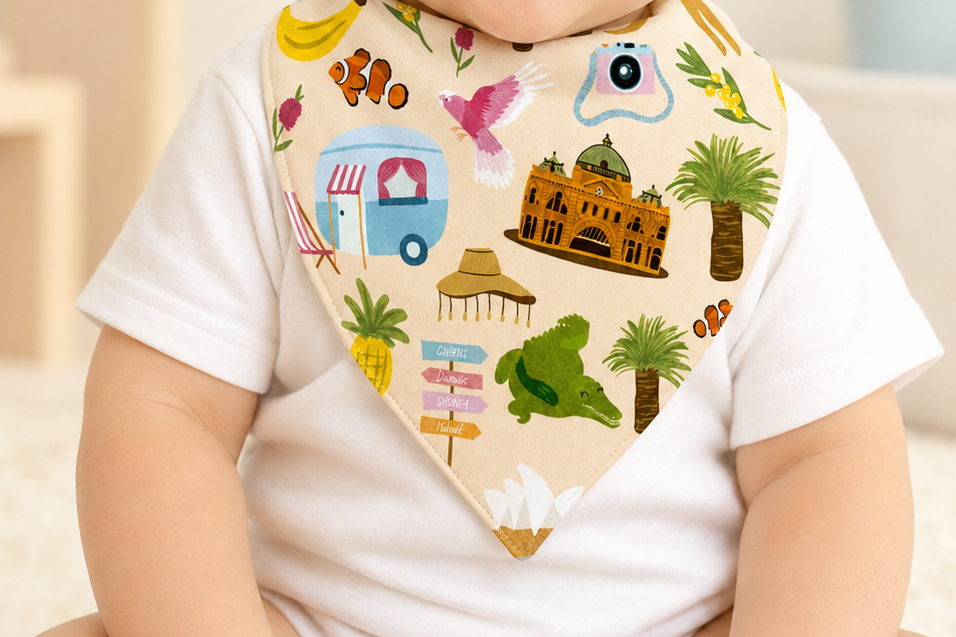 A smiling baby wearing a white short-sleeve bodysuit and a beige bib with colorful travel-themed illustrations, sitting in a softly lit room.