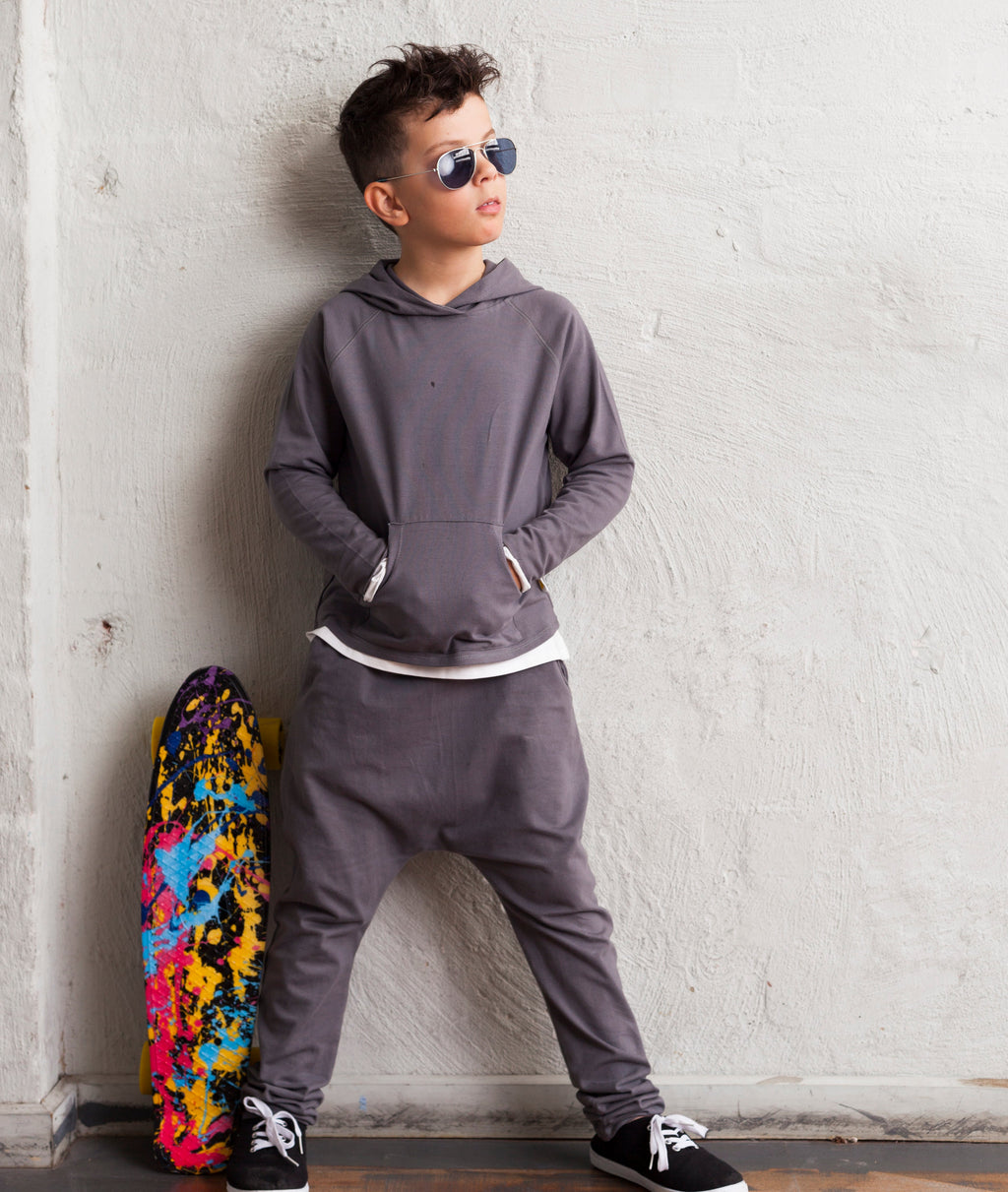 Person wearing a gray hoodie and pants leaning against a white brick wall with a colorful skateboard on the ground.