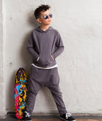 Person wearing a gray hoodie and pants leaning against a white brick wall with a colorful skateboard on the ground.