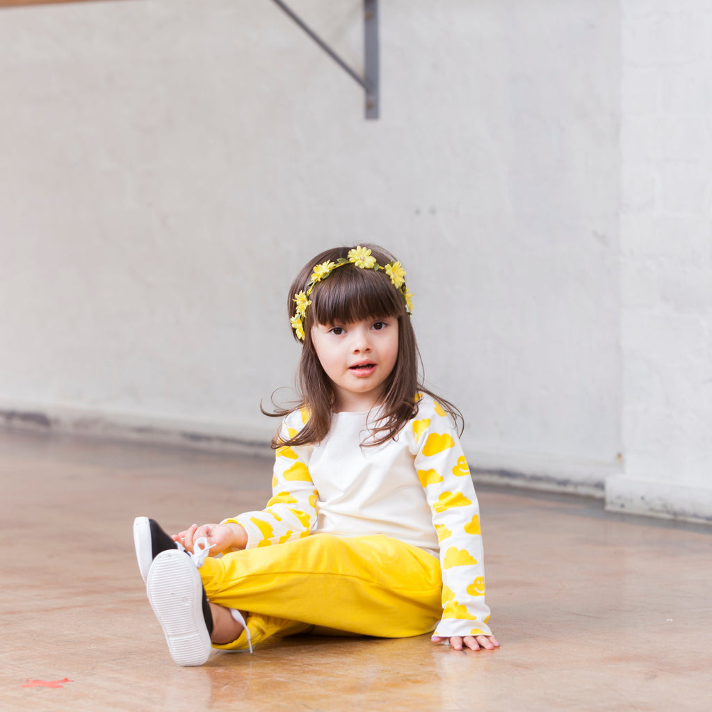 Child sitting on the floor in a dance studio wearing a yellow and white outfit with a floral headband.