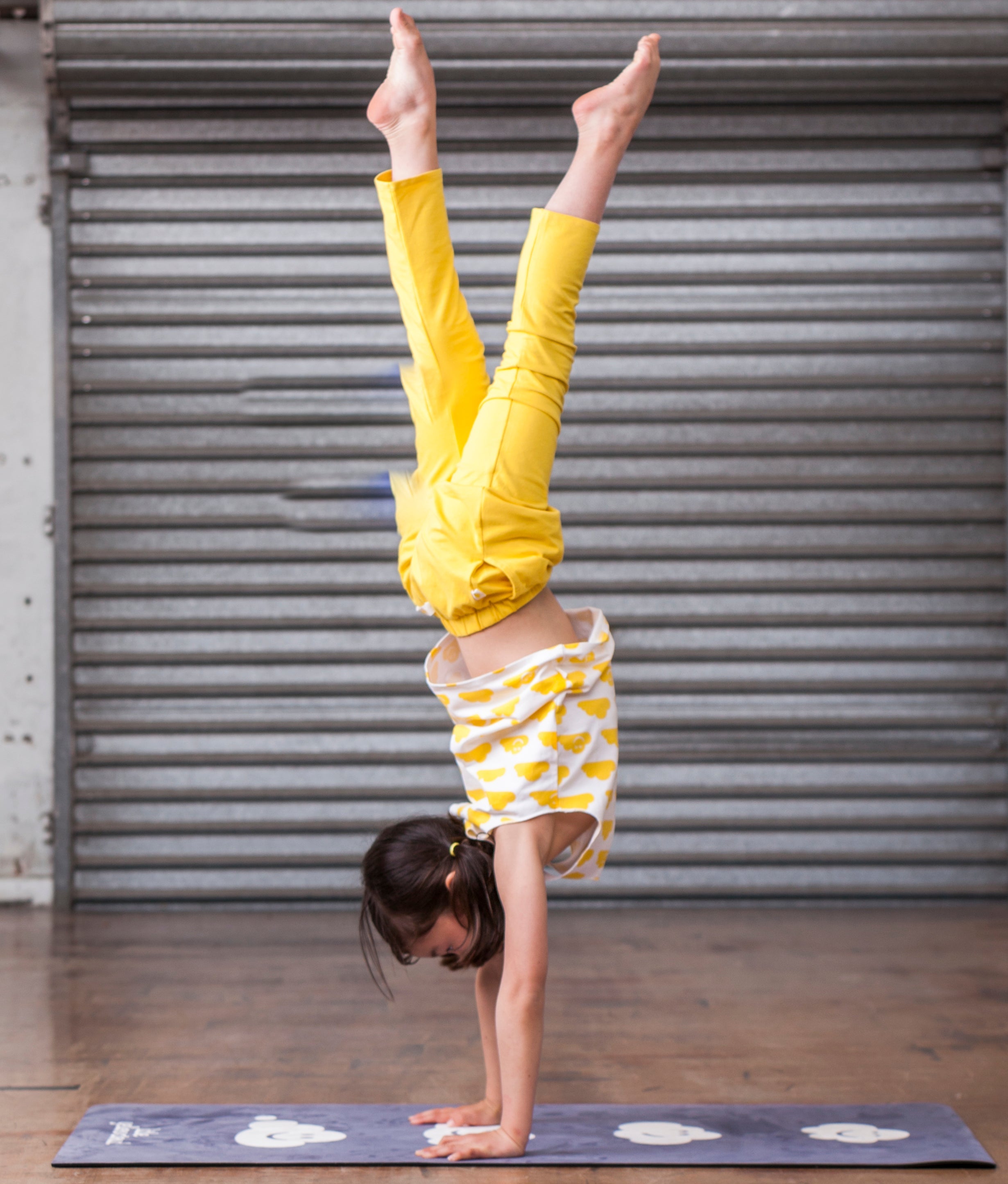 Child performing a handstand on a mat in front of a gray metal shutter.