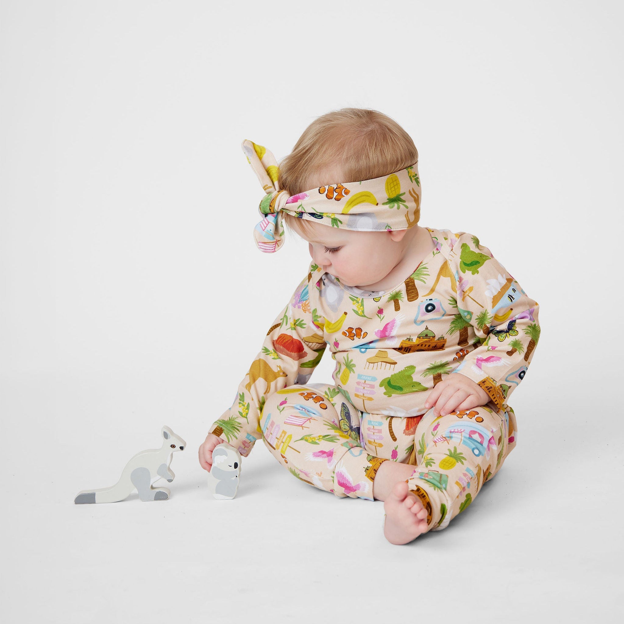 Baby girl wearing Aussie Road Trip organic cotton outfit with matching knot headband sitting beside wooden kangaroo toys