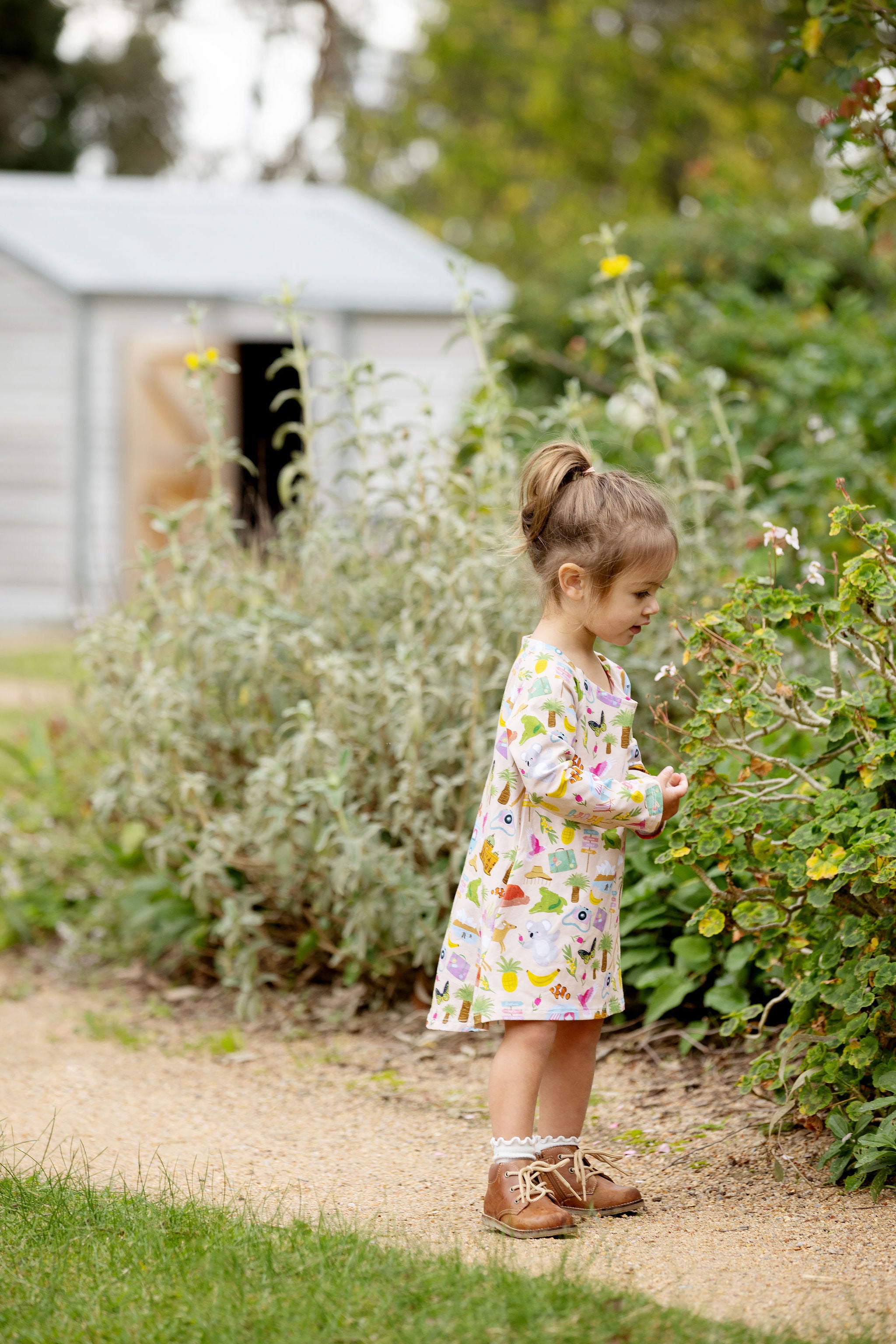 Little girl wearing Pebble and Poppet Aussie Road Trip Organic Cotton Dress standing in an Australian garden featuring kangaroos, koalas, caravans and travel-inspired illustrations.