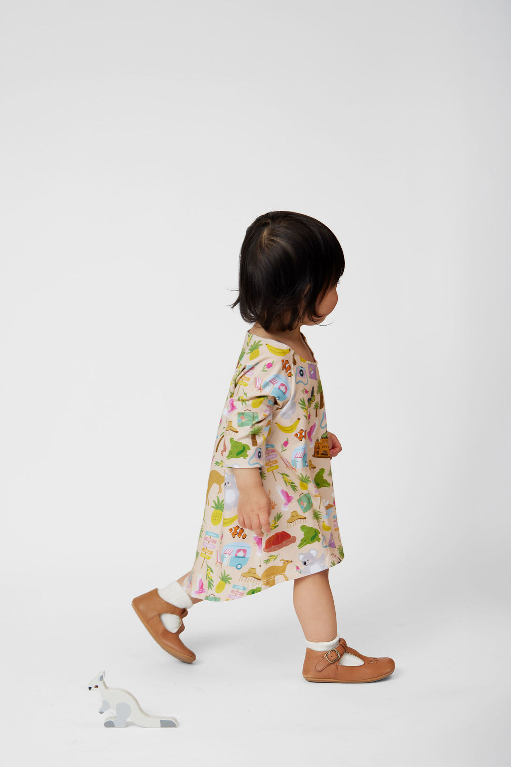 Side profile of a toddler wearing the Aussie Road Trip Organic Cotton Dress featuring Australian animals, caravans, tropical motifs and family holiday illustrations on a soft blush background.