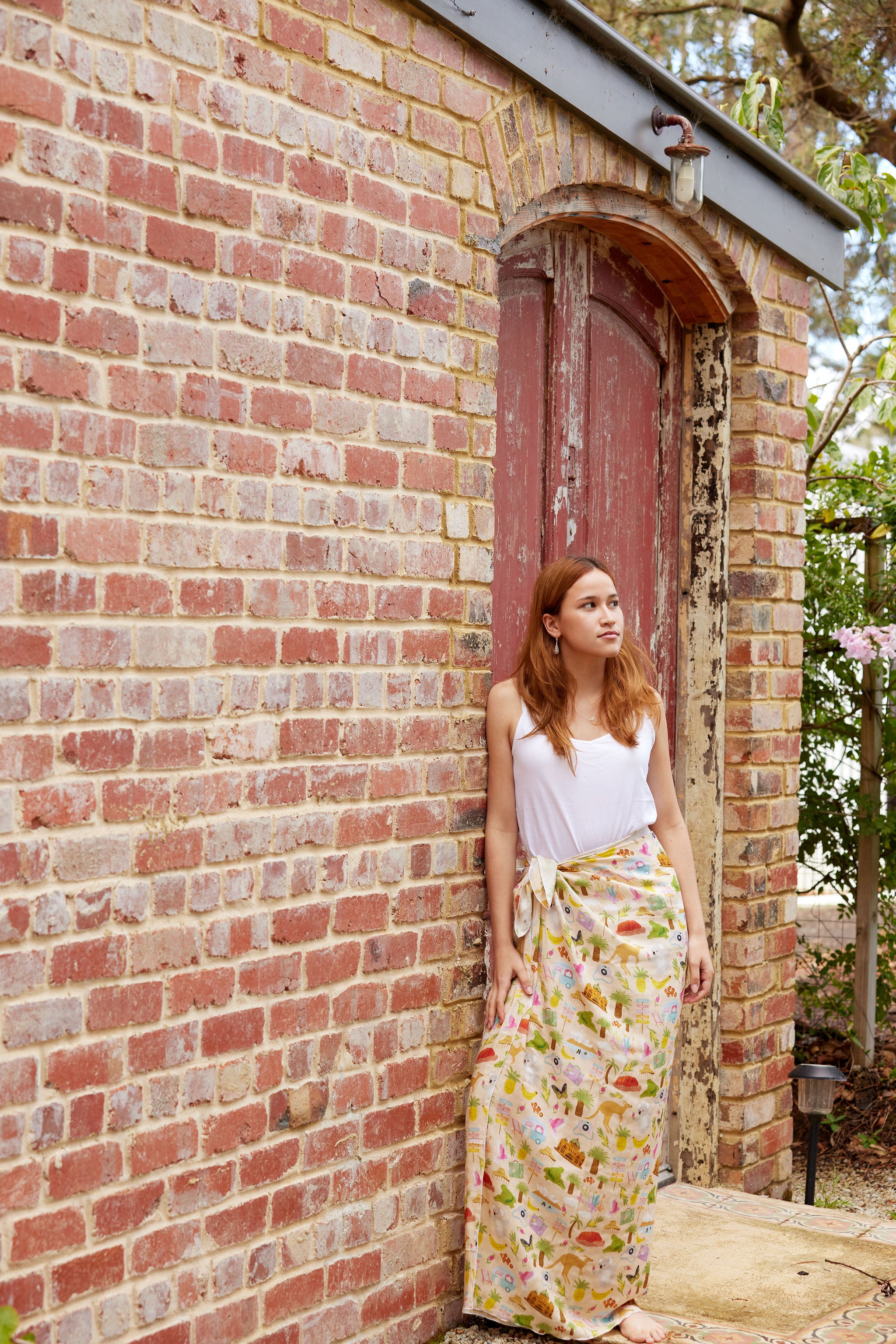 Young woman wearing the Aussie Road Trip print sarong by Christie Williams leaning against a rustic brick wall outdoors