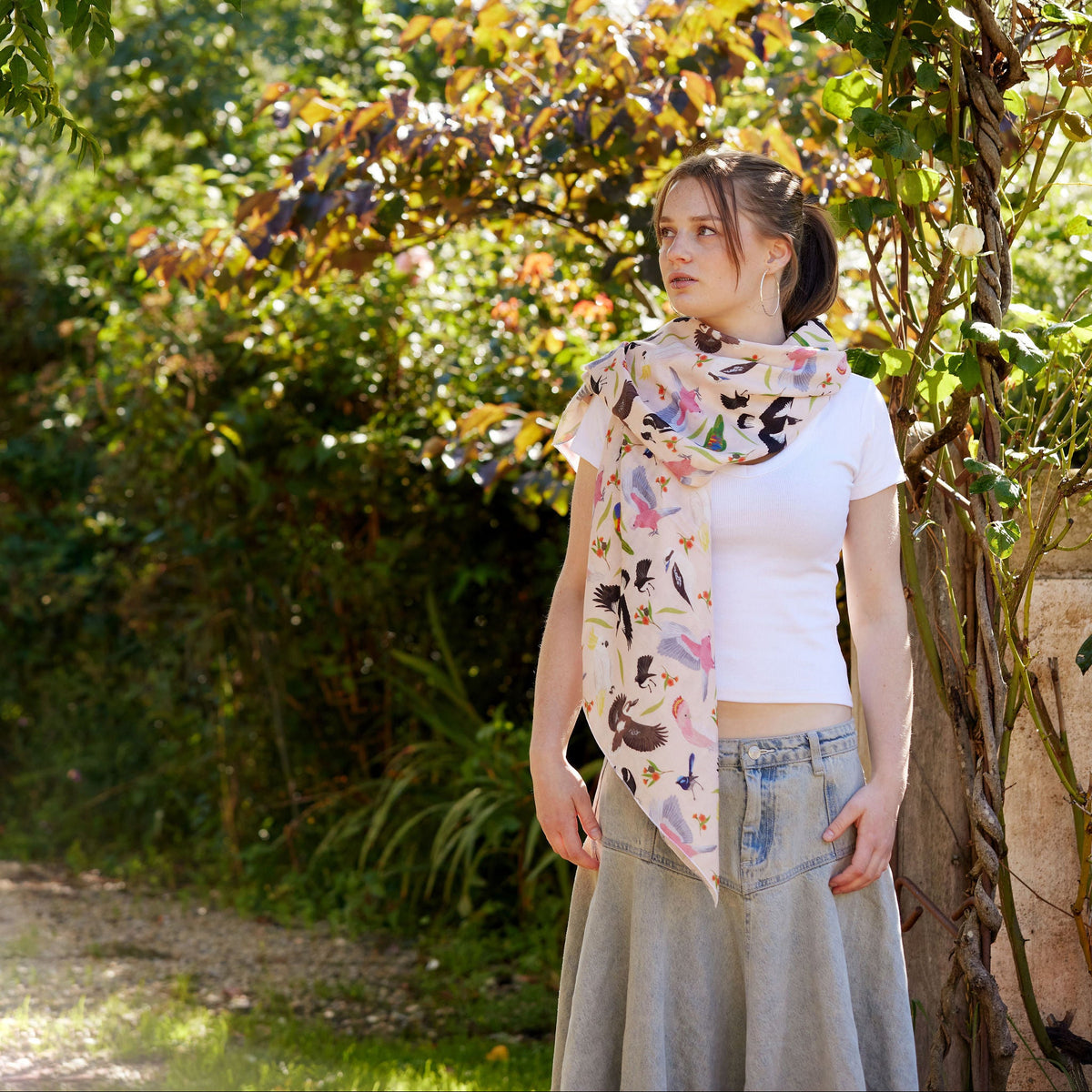 Young woman wearing a soft Australian bird print scarf standing beneath a leafy garden arch in warm sunlight