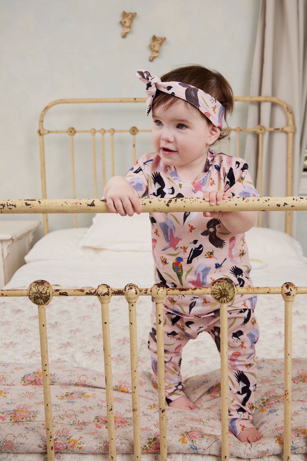 Baby standing in vintage iron bed wearing Backyard Birds baby pants and matching Australian bird print top with knotted headband