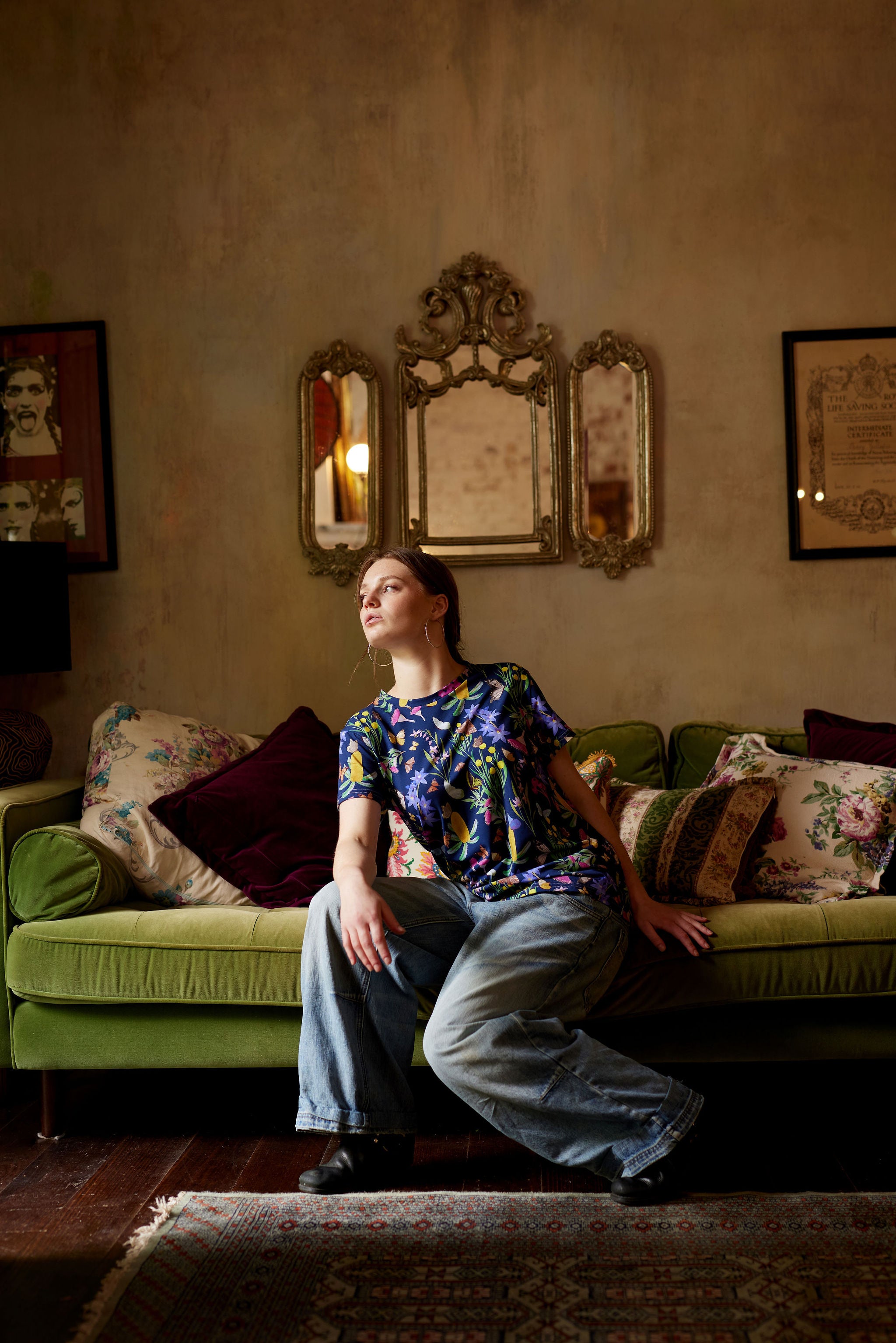 Model wearing Butterfly Flora organic cotton t-shirt sitting on green velvet couch in moody vintage interior.