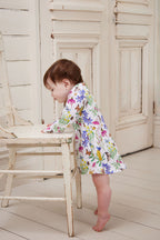 Baby girl standing beside a vintage chair wearing Butterfly Flora long sleeve dress indoors.