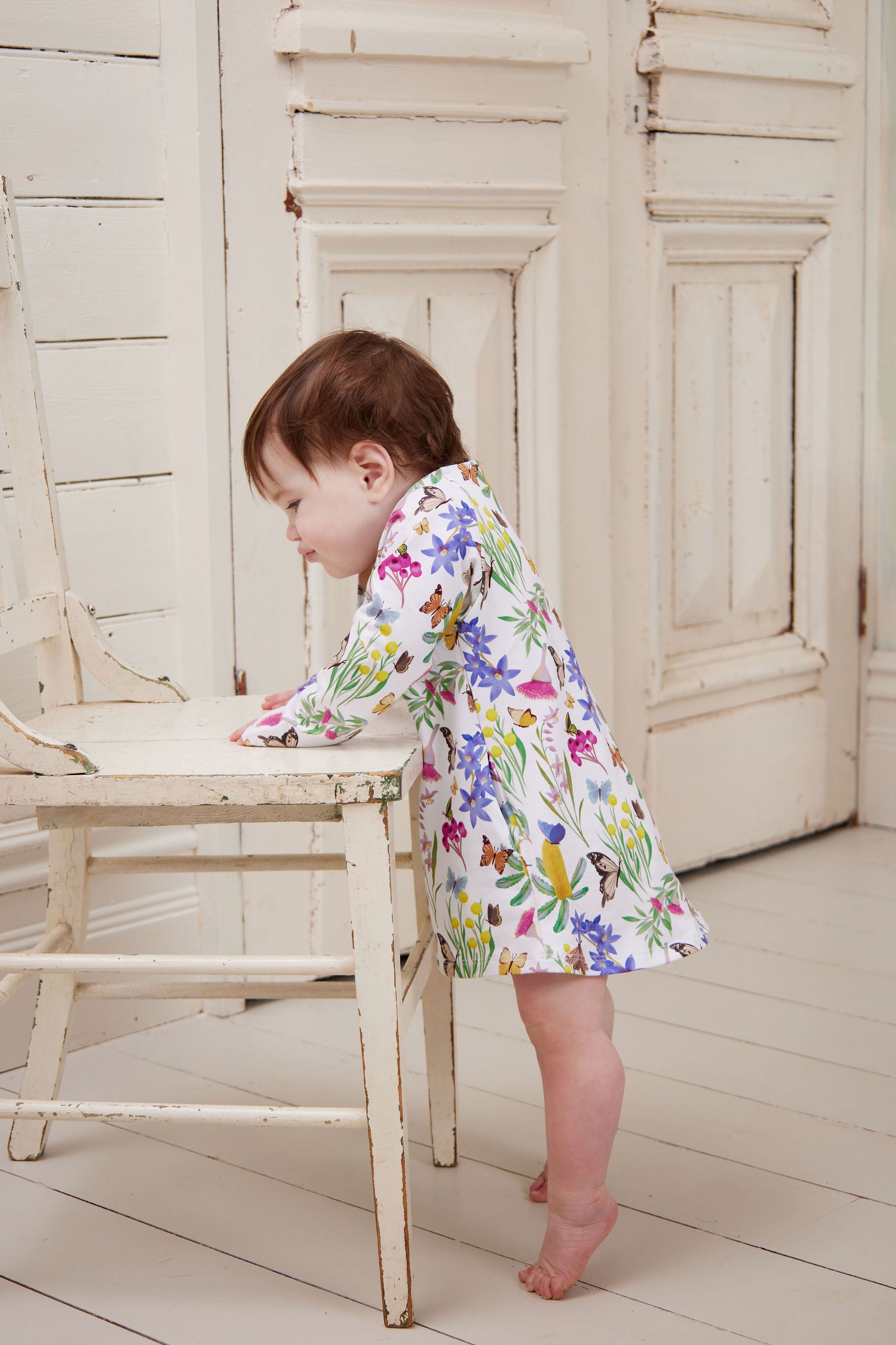 Baby girl standing beside a vintage chair wearing Butterfly Flora long sleeve dress indoors.