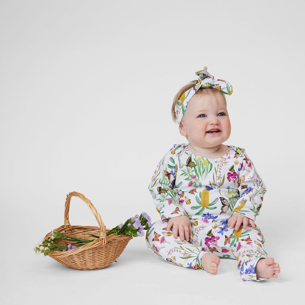Baby girl wearing Pebble and Poppet Butterfly Flora romper with matching floral headband sitting beside a basket of flowers.
