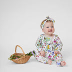 Baby girl wearing Pebble and Poppet Butterfly Flora romper with matching floral headband sitting beside a basket of flowers.