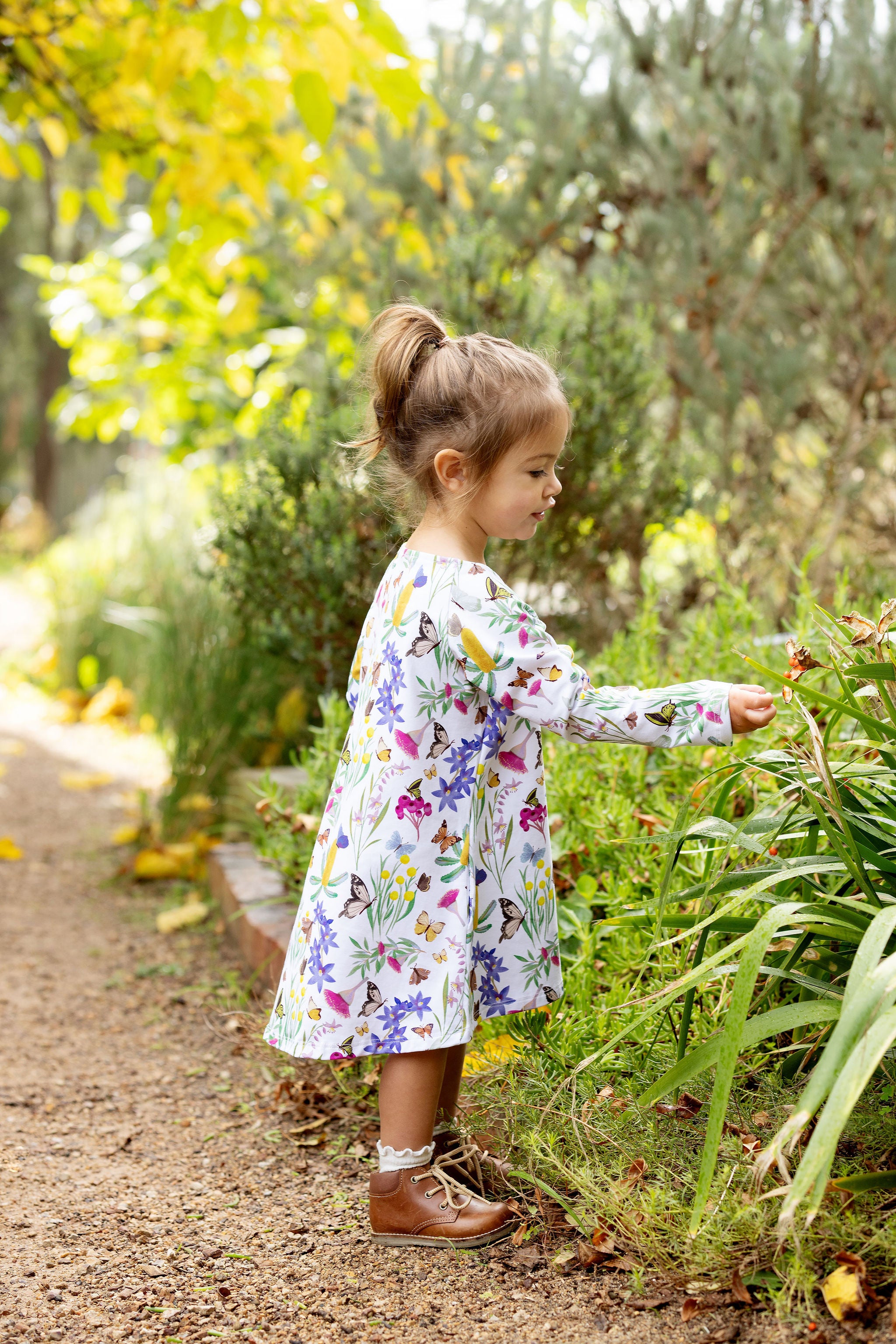 Little girl wearing Butterfly Flora long sleeve dress standing in a garden surrounded by greenery and flowers.