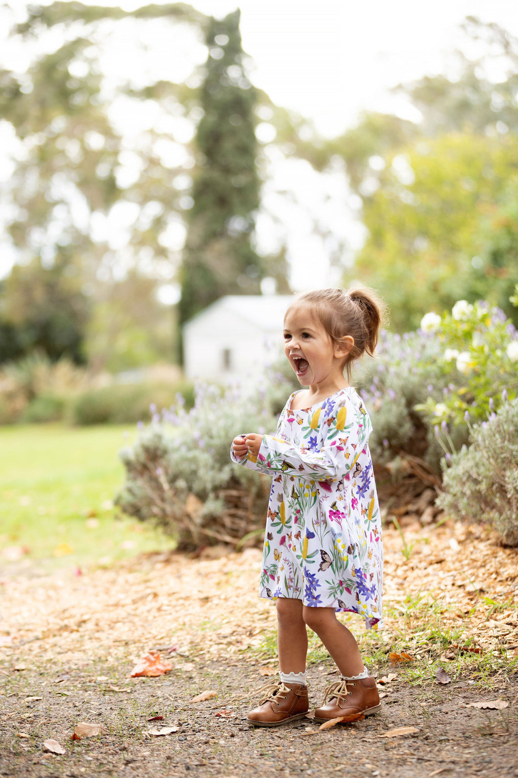 Little girl wearing Butterfly Flora long sleeve dress standing in a garden surrounded by greenery and flowers.