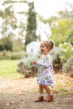Little girl wearing Butterfly Flora long sleeve dress standing in a garden surrounded by greenery and flowers.