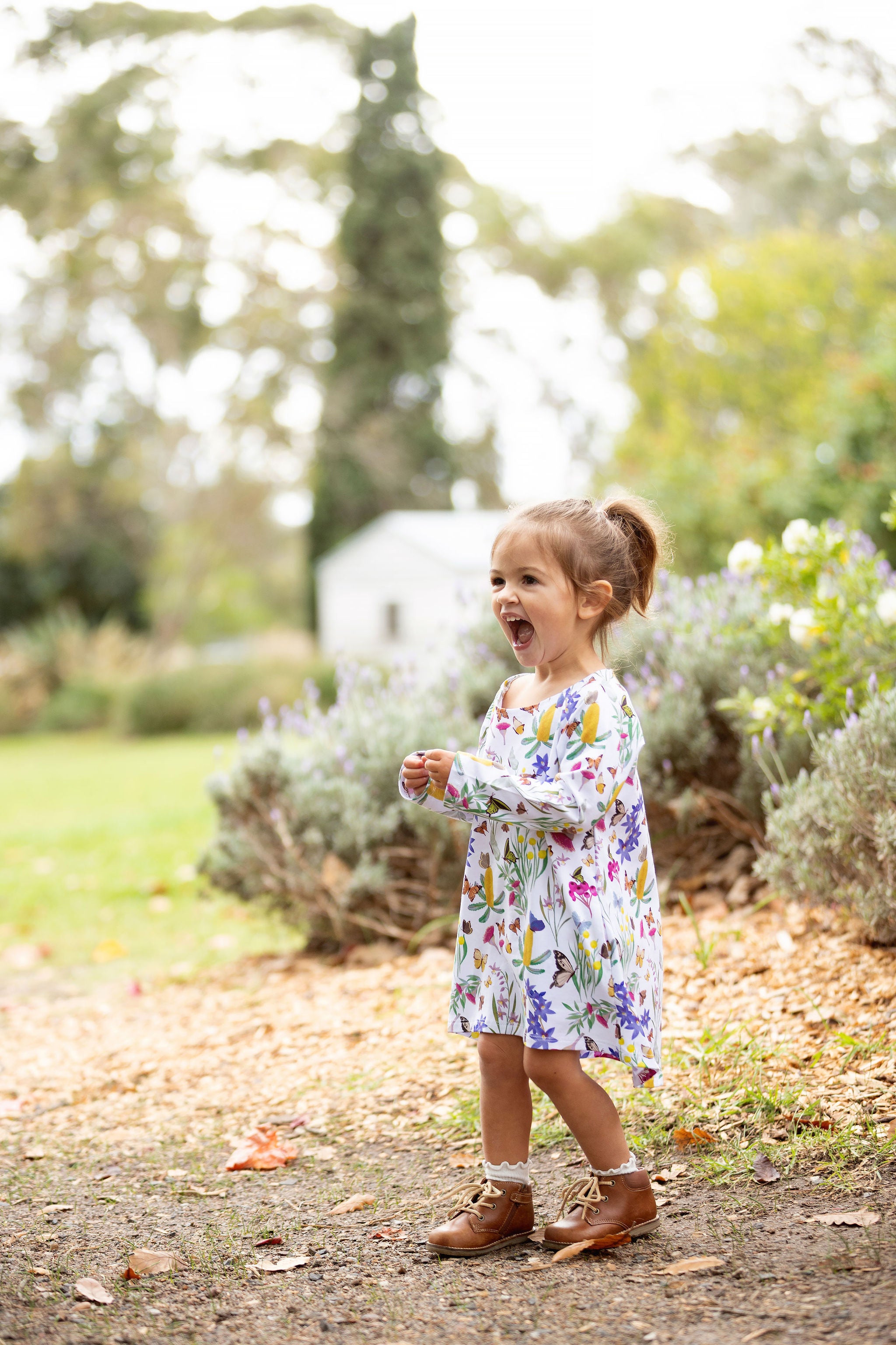 Little girl wearing Butterfly Flora long sleeve dress standing in a garden surrounded by greenery and flowers.