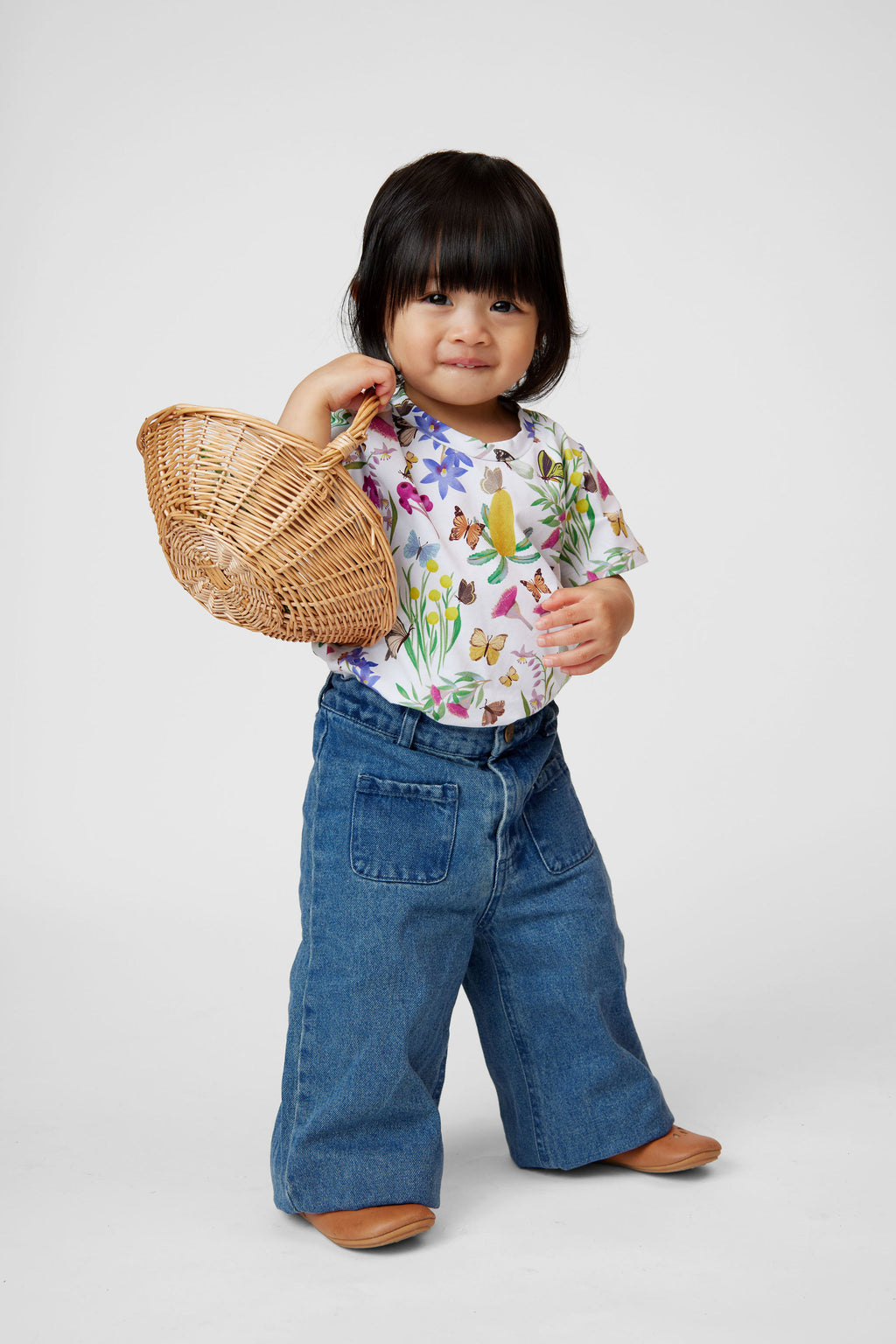 Little girl wearing Butterfly Flora organic cotton t-shirt with denim pants holding a wicker basket in a studio setting.