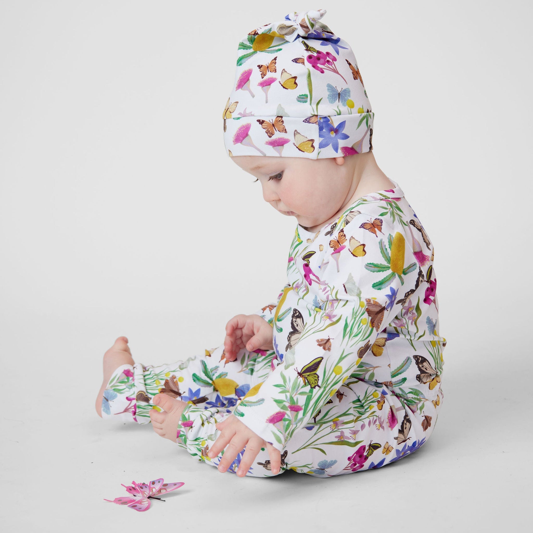 Baby wearing Butterfly Flora Australian made organic cotton romper and matching knot beanie sitting beside a butterfly toy.