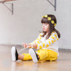 Child sitting on the floor wearing a yellow and white outfit with flower headband.