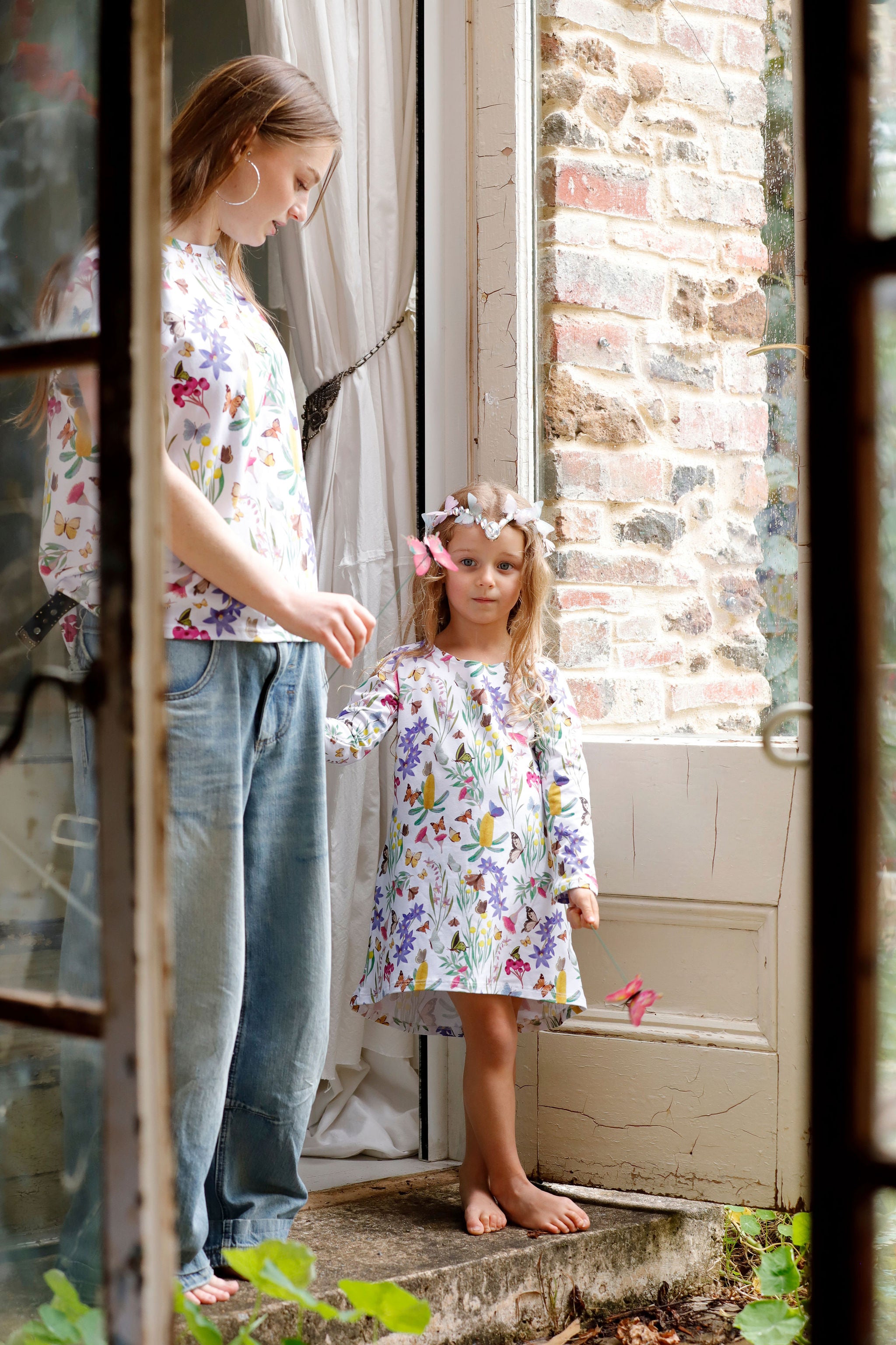 Mother and child wearing matching Butterfly Flora floral butterfly outfits beside rustic cottage doorway.