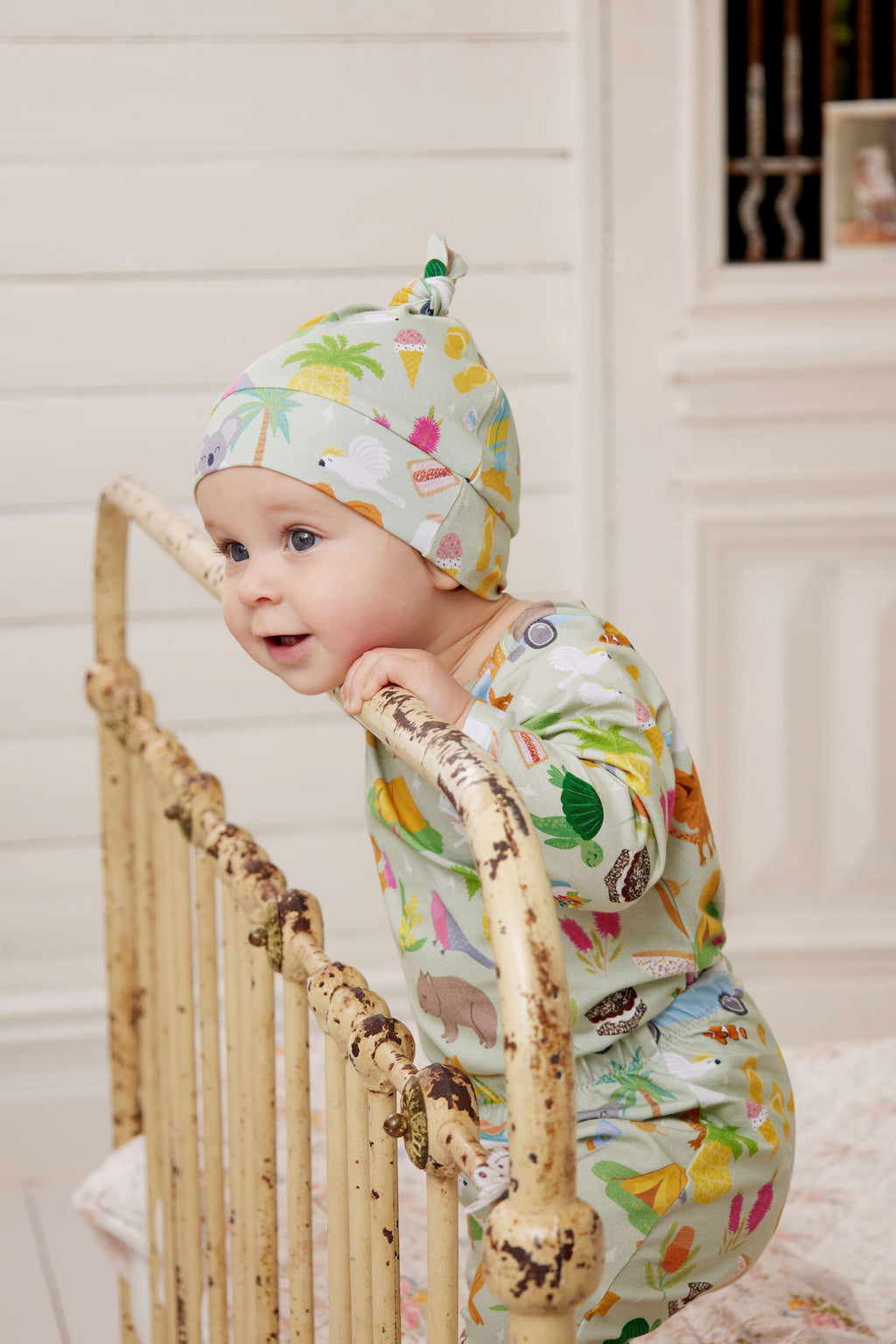 Baby wearing Summer Down Under organic cotton beanie and matching outfit standing indoors featuring whimsical Australian print with kangaroos, kombis and camping icons. Designed and made in Australia.