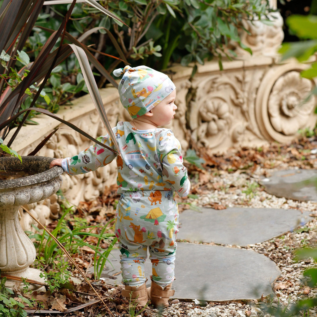 Baby wearing Pebble and Poppet Summer Down Under organic cotton outfit and matching knot beanie while holding flowers in a cottage garden setting. Features whimsical Australian print with native animals, kombis and camping motifs. Designed and made in Australia.
