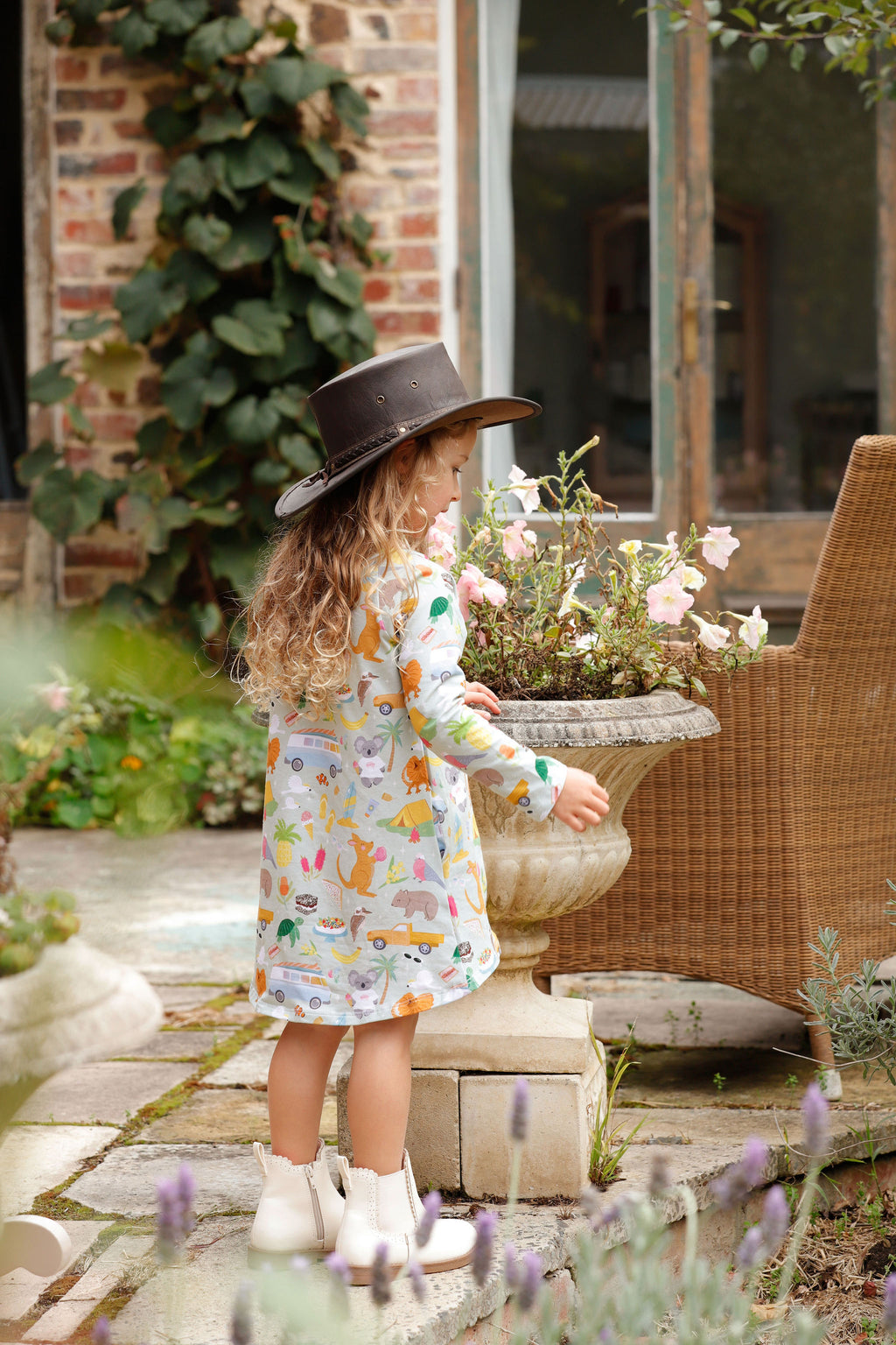 Little girl wearing Pebble and Poppet Summer Down Under organic cotton dress in Australian inspired print standing in cottage garden wearing brown outback hat and white boots. Designed and made in Australia.