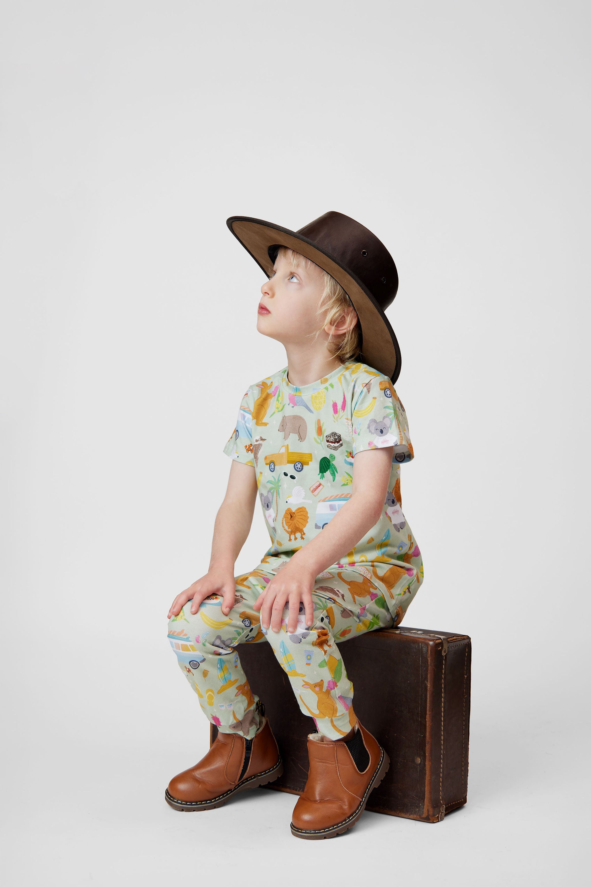 Child wearing Summer Down Under organic cotton kids outfit by Pebble and Poppet sitting on vintage suitcase wearing wide brim Australian outback hat. Designed and made in Australia.