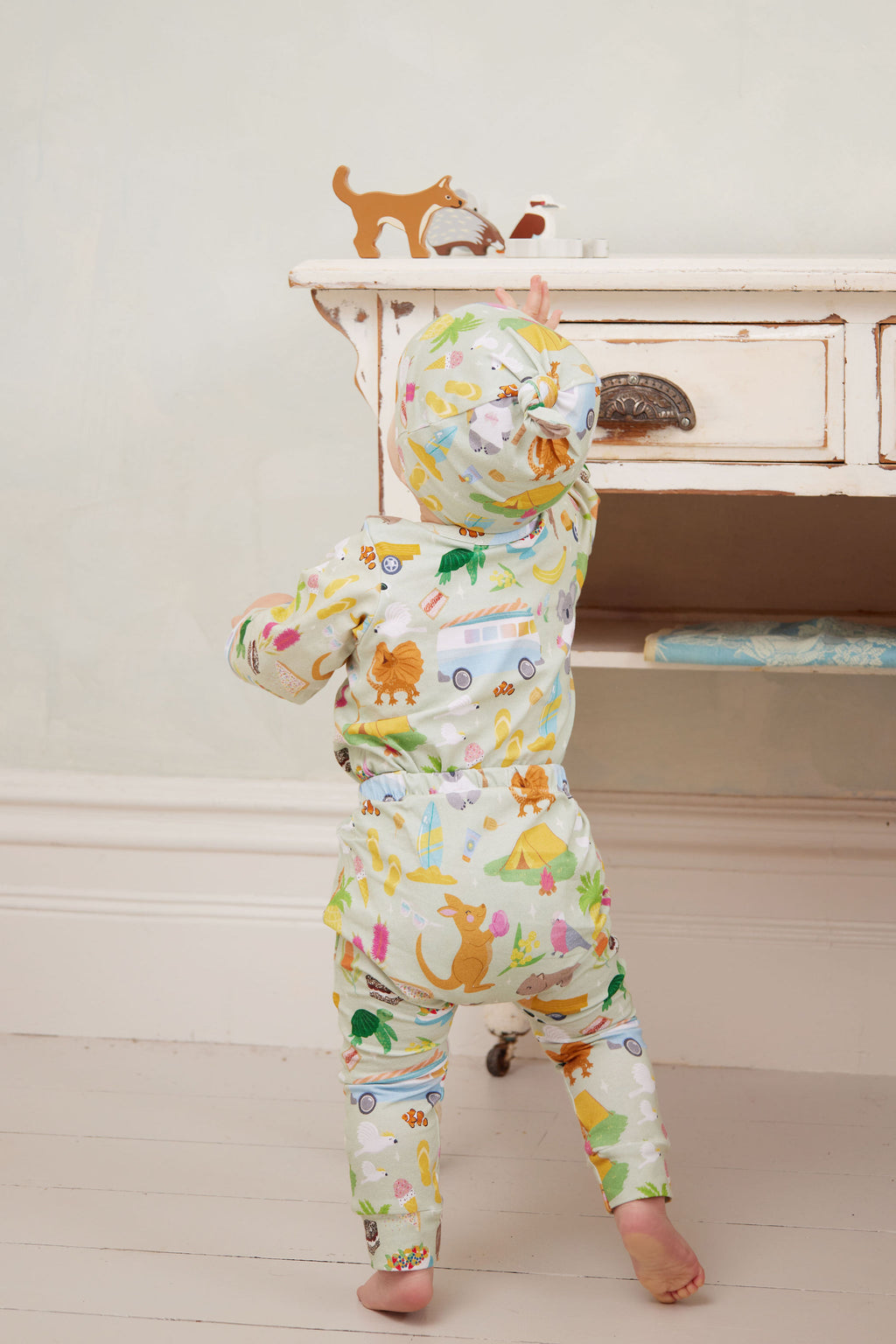Baby wearing Pebble and Poppet Summer Down Under organic cotton beanie and outfit leaning over vintage cot in soft neutral nursery. Designed and made in Australia.