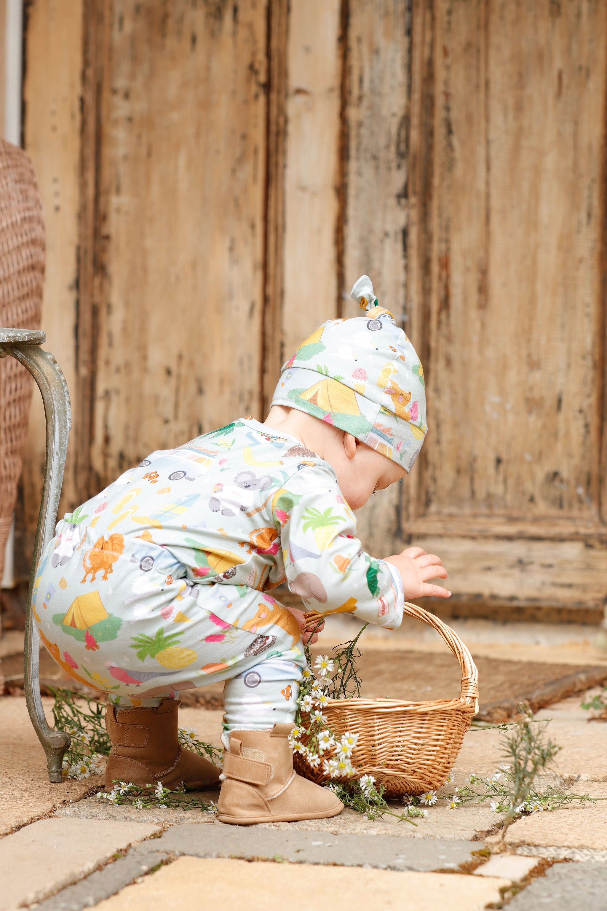 Baby wearing Pebble and Poppet Summer Down Under organic cotton pants and matching knot beanie while crouching beside a basket of flowers outdoors. Features whimsical Australian print with kombis, kangaroos and camping icons. Designed and made in Australia