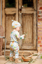 Baby wearing Pebble and Poppet Summer Down Under organic cotton romper and matching knot beanie while holding flowers beside a basket in a cottage garden. Features whimsical Australian print with kombis, camping, native animals and summer holiday motifs. Designed and made in Australia