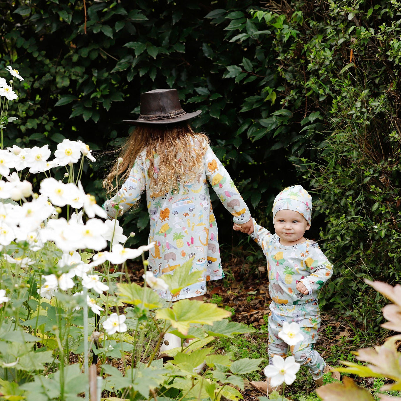 Children wearing matching Summer Down Under organic cotton outfits by Pebble and Poppet walking through garden surrounded by flowers and greenery. Designed and made in Australia.